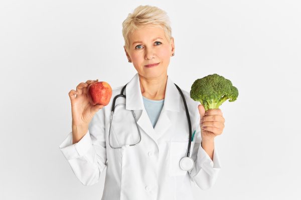 Attractive confident mature Caucasian female doctor holding red apple and green broccoli as part of healthy diet to reduce risk of some chronic diseases. Food, nutrition and health concept